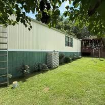 A long, horizontal mobile home with cream siding above a green base, set in a grassy yard under a tree canopy.
