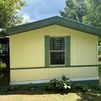 A yellow manufactured home with a green roof, shutters, and foundation, set against a background of trees.