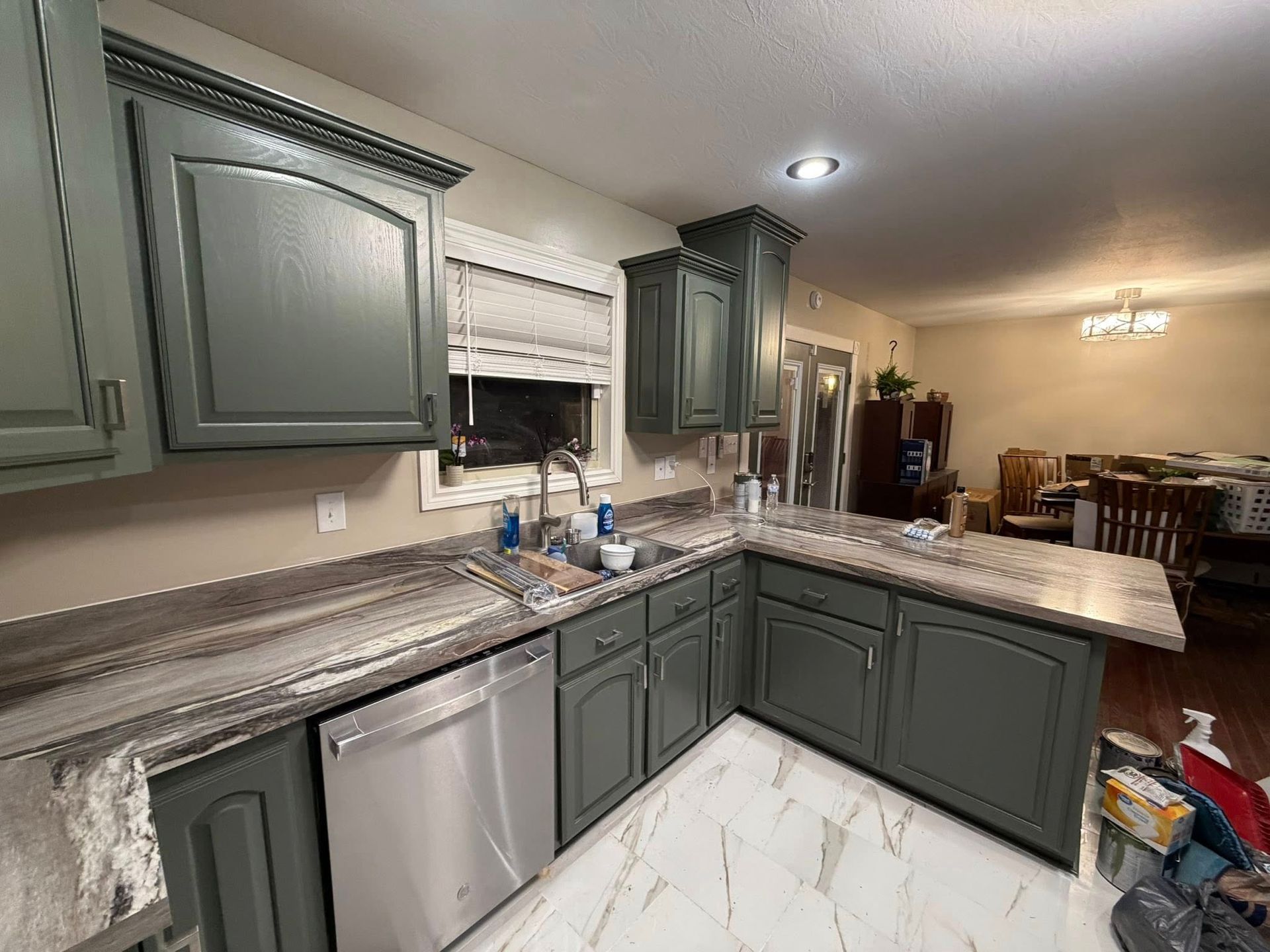 Kitchen with dark gray cabinets, marble-patterned countertops, stainless steel dishwasher, and white tile floors.