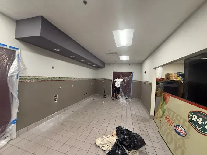 A person in a room undergoing renovation with gray walls, tiled floors, a dark soffit, and plastic-covered door frames.