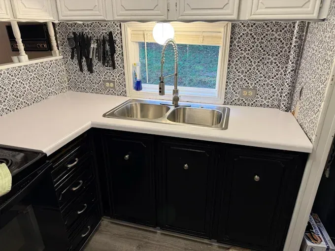 A kitchen sink area with a silver faucet, white countertops, black cabinets, and patterned black-and-white wallpaper.
