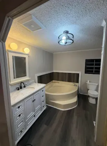 Modern bathroom with a white vanity, a corner garden tub, wood-look flooring, a mirror, and a toilet against a white wall.