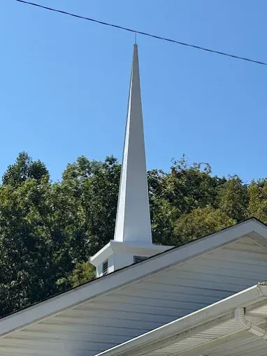 A tall, white steeple rises above the roofline of a building, set against a background of green trees and a blue sky.