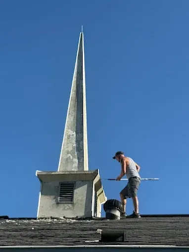A person stands on a shingled roof, holding a long pole near a white church spire against a clear blue sky.