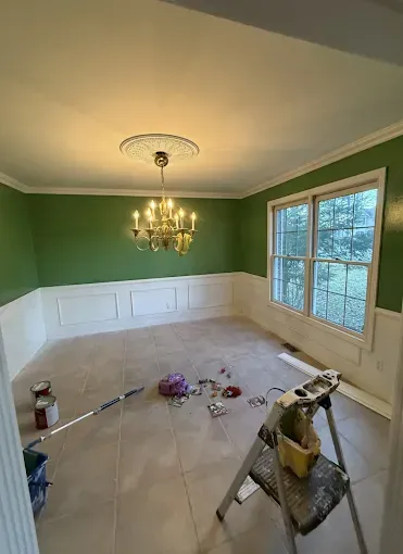 A dining room undergoing renovation with green walls, white wainscoting, a chandelier, and painting supplies on the floor.