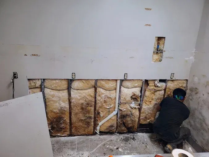 A worker repairs drywall in a room with exposed wall studs and insulation.