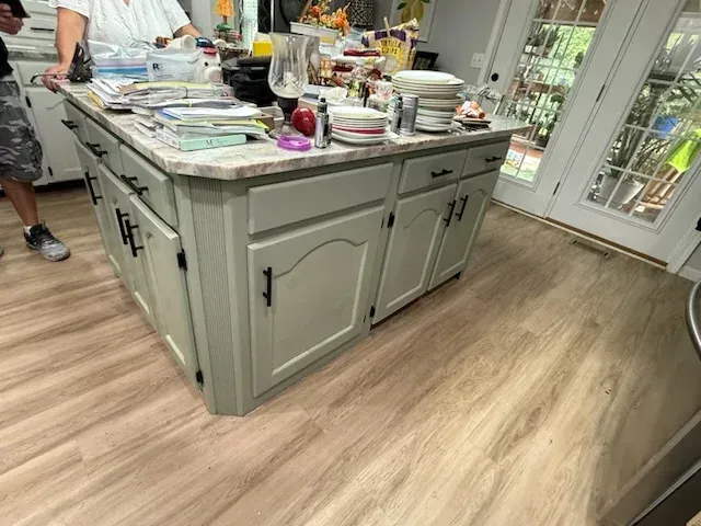A kitchen island with light green cabinets and black hardware sits on light wood-look flooring in a brightly lit room.