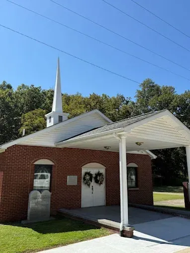 A small brick church with a white steeple and a covered entryway, set against a backdrop of green trees under a blue sky.
