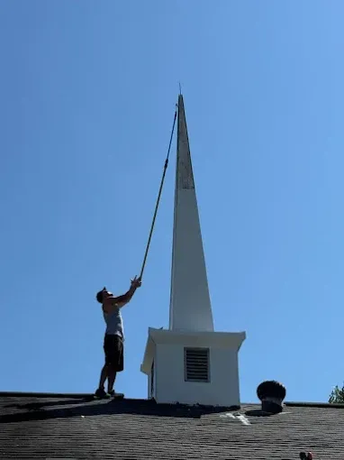 A person on a dark roof uses a long pole to reach the top of a white church steeple against a clear blue sky.