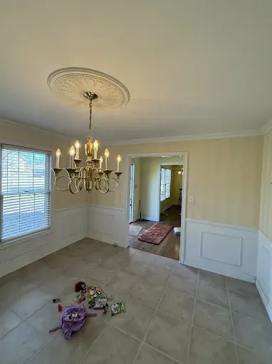 A dining room with tile floors, yellow striped walls, white wainscoting, a gold chandelier, and a pile of toys on the floor.