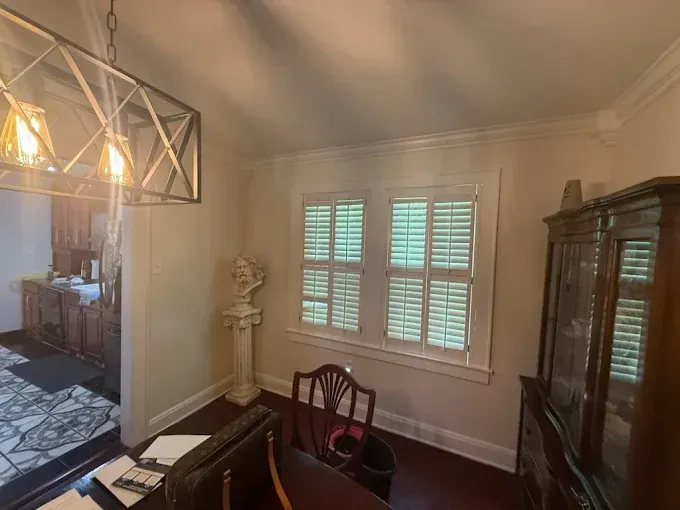 A dining area with a modern chandelier, white walls, shuttered windows, a pedestal statue, and a wooden cabinet.