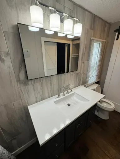 A bathroom vanity with a rectangular mirror, a white sink, dark cabinets, and four cylinder lights on a tiled wall.