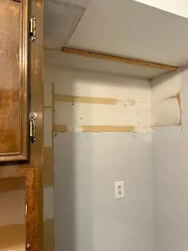 A section of a wall in a home with exposed drywall seams, tape, and a wood strip where cabinets were removed.
