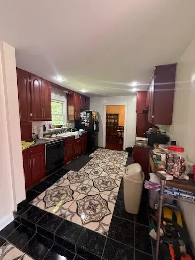 A kitchen featuring dark wood cabinets, a patterned rug down the center, black floor tiles, and a doorway to another room.