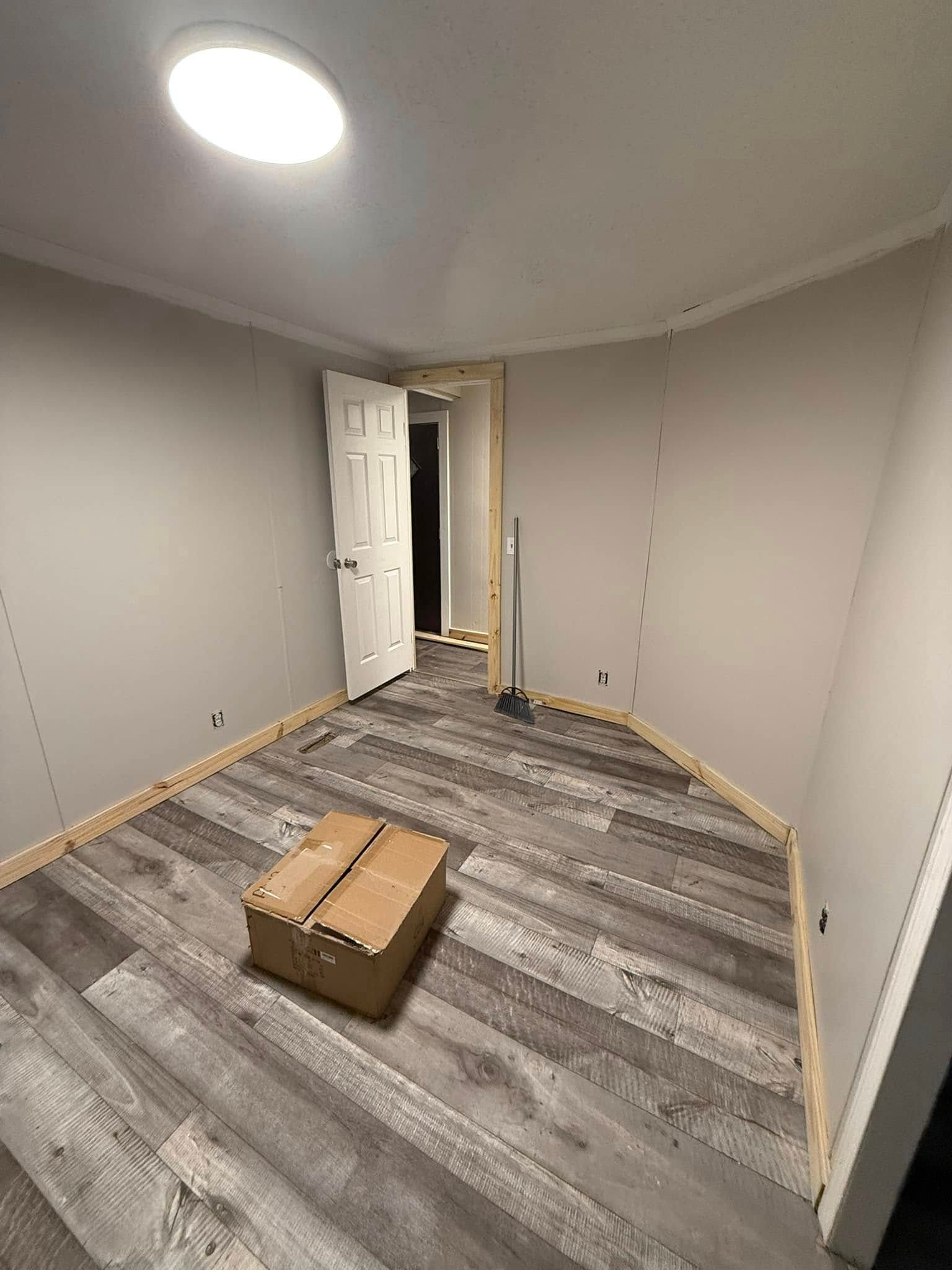 A room with grey walls, light wood-look vinyl flooring, unpainted baseboards, a white door, and a brown box in the center.