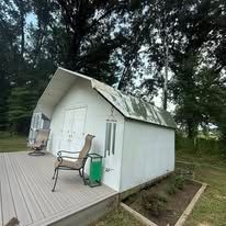 A white shed with a deck, a brown chair, and a wind chime, set outdoors near a wooded area.