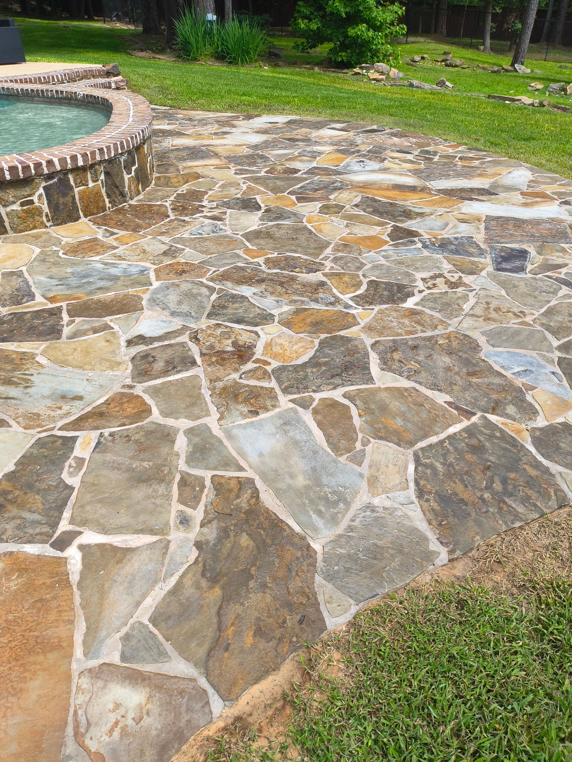 Stone patio beside a pool. Flagstone paving with various brown, grey, and tan stones. Green grass surrounds.