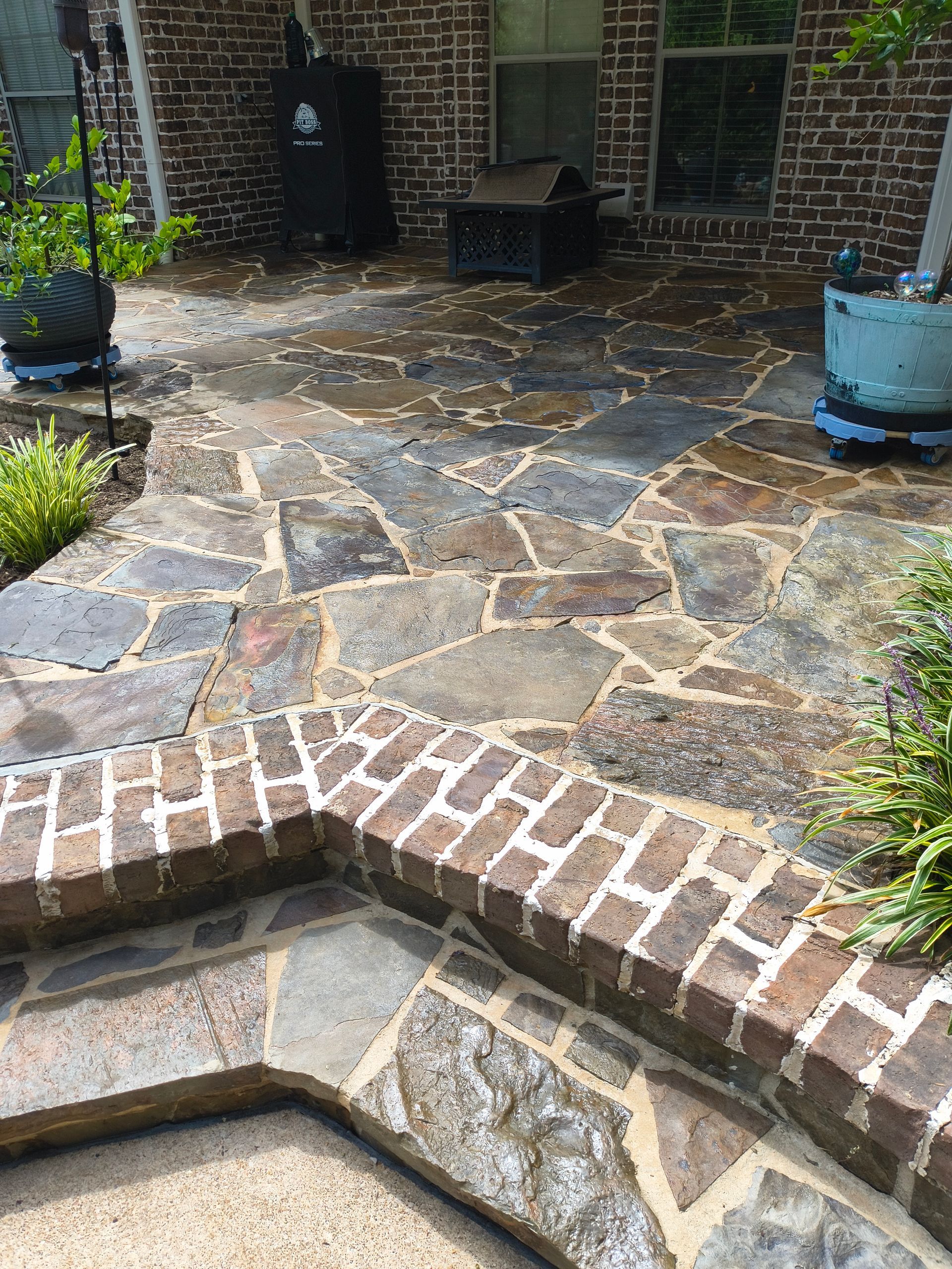 Stone patio with brick trim, steps leading up to a house with a smoker and potted plants.