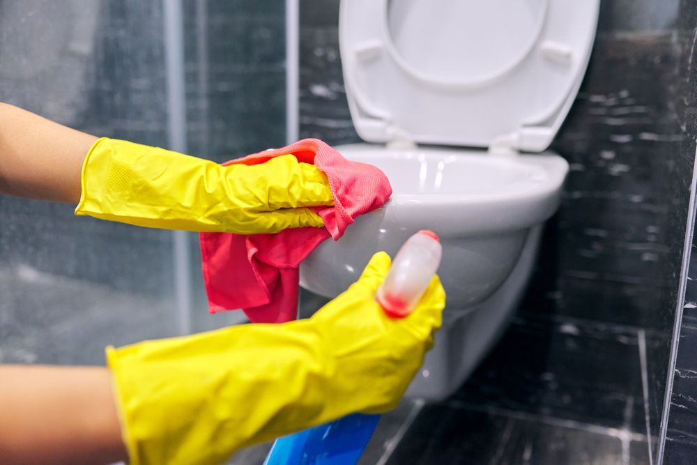 Person wearing yellow gloves sprays and wipes a toilet in a bathroom with a red cloth.