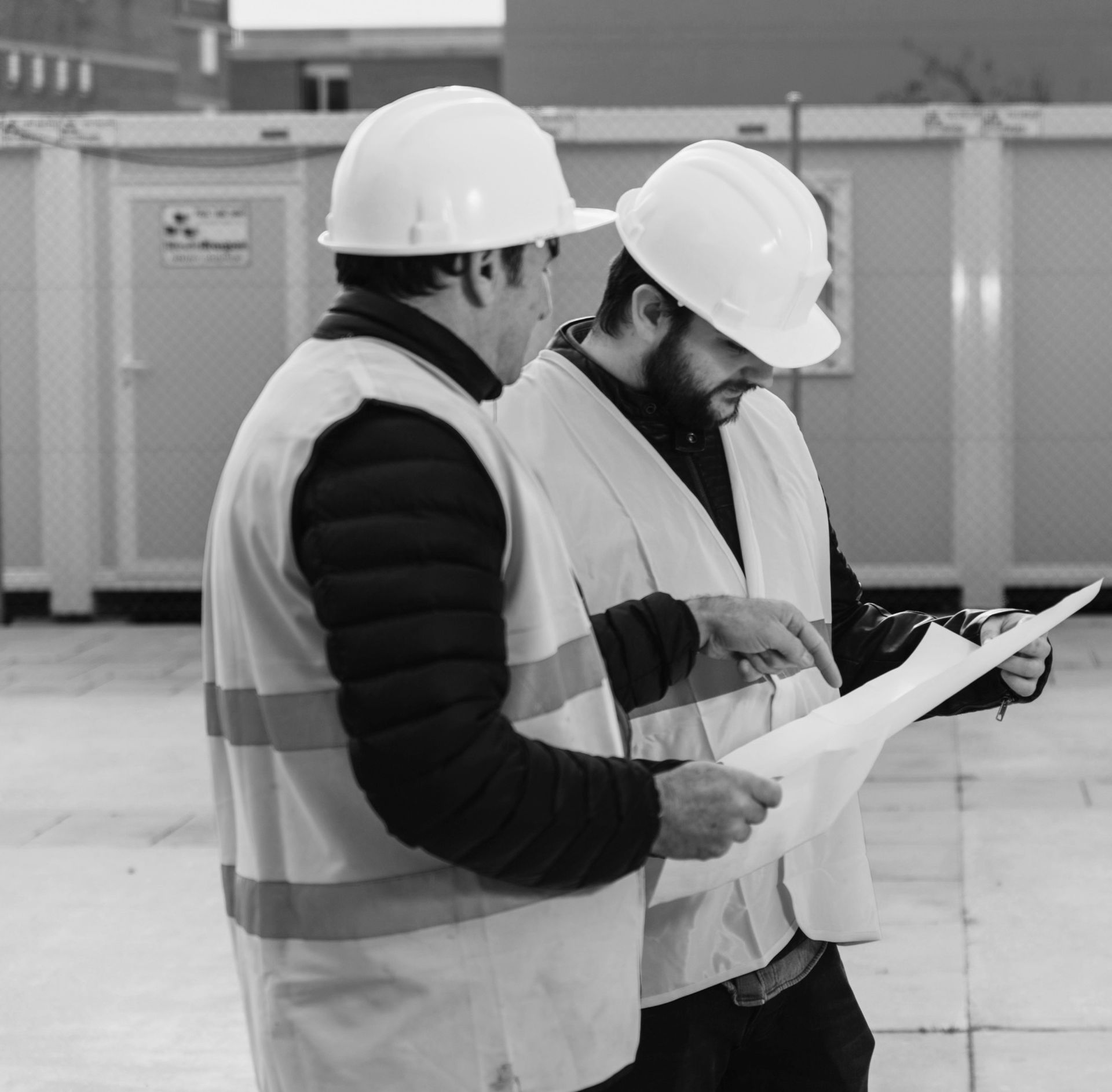 Two construction supervisors wearing white hard hats and high-visibility safety vests conferring over a site blueprint, illustrating how group captives align members based on shared operational methods and safety protocols.