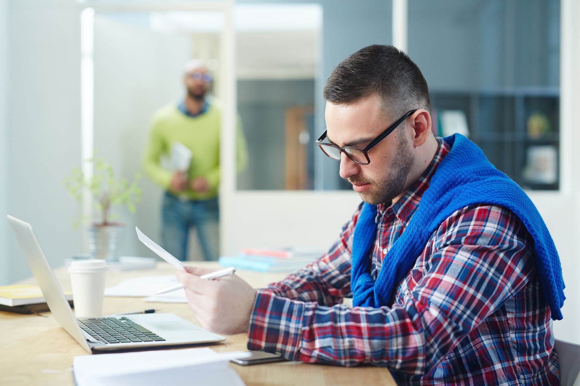 A focused professional in a plaid shirt and glasses sits at a desk, carefully reviewing a physical document while working on a laptop, representing the financial maturity and detailed documentation required in group captive insurance.