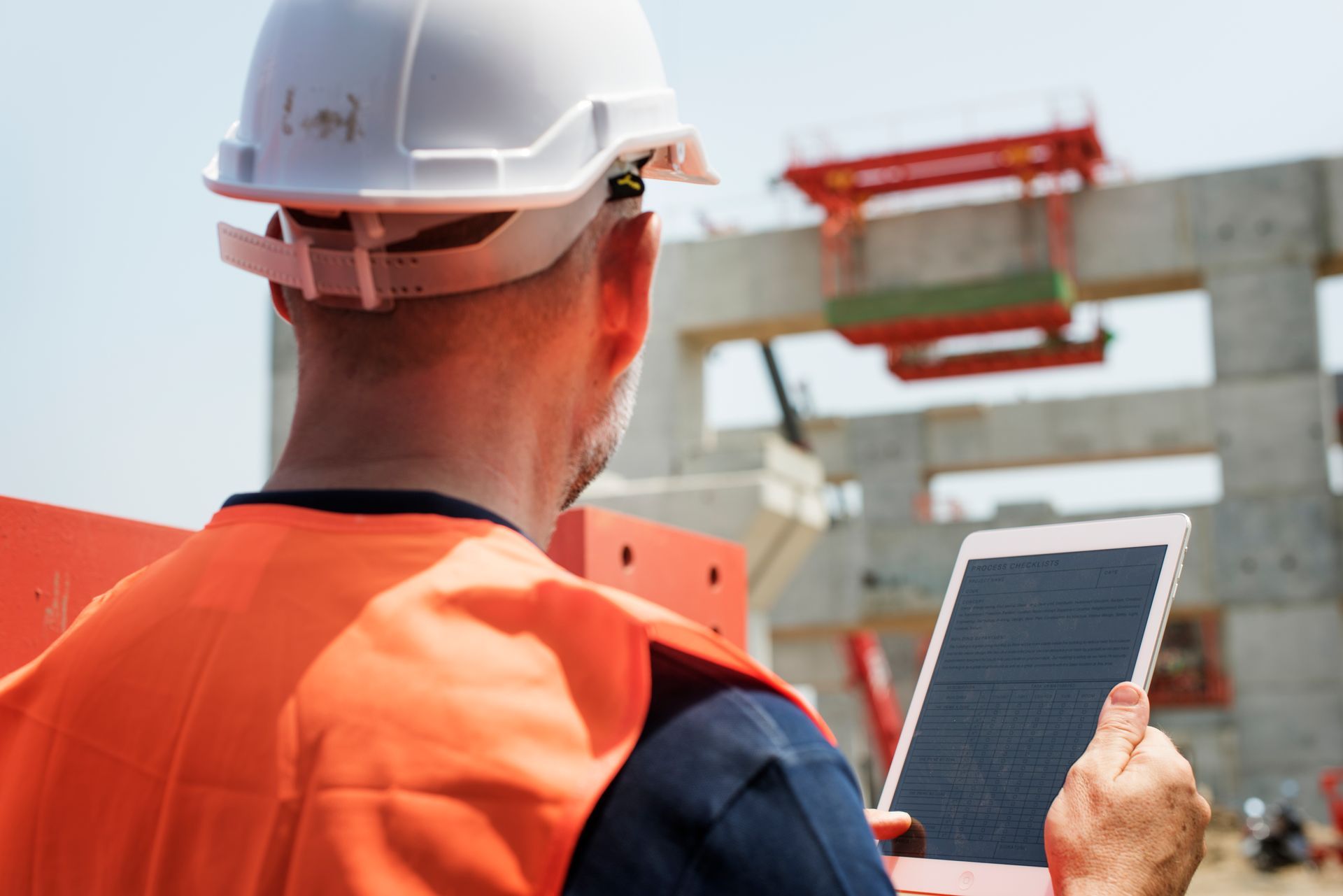 Over-the-shoulder view of a construction supervisor in a white hard hat and orange safety vest using a tablet to monitor a job site, representing the active oversight and technological readiness required for captive qualification.