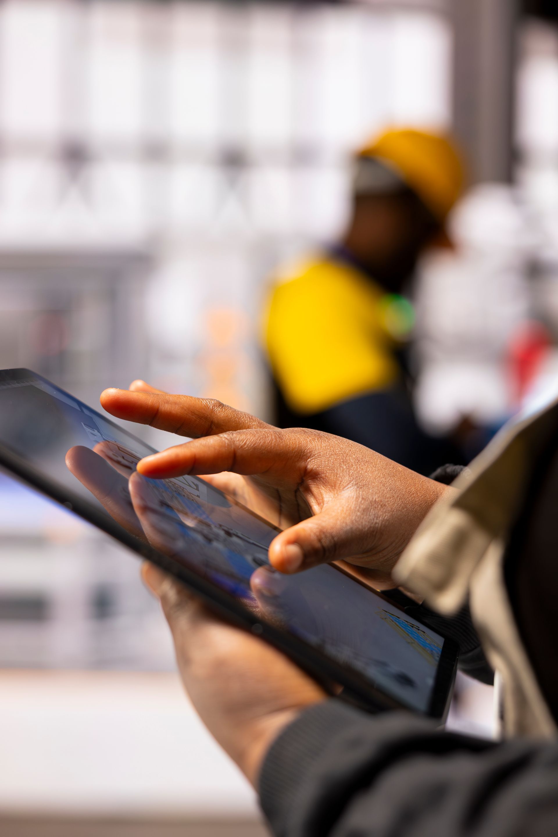 A close-up of hands using a digital tablet to review site data on a construction project, with a blurred worker in a high-visibility yellow vest in the background, illustrating a hands-on, operator-focused assessment.