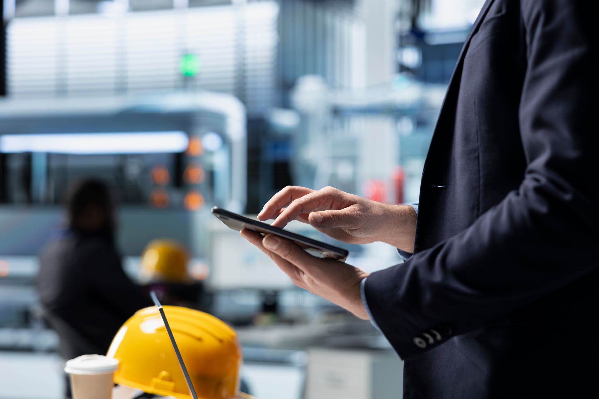 A close-up of a professional in a dark suit using a digital tablet in a modern, high-tech industrial environment with a yellow hard hat in the foreground, representing the intersection of field operations and sophisticated financial management.