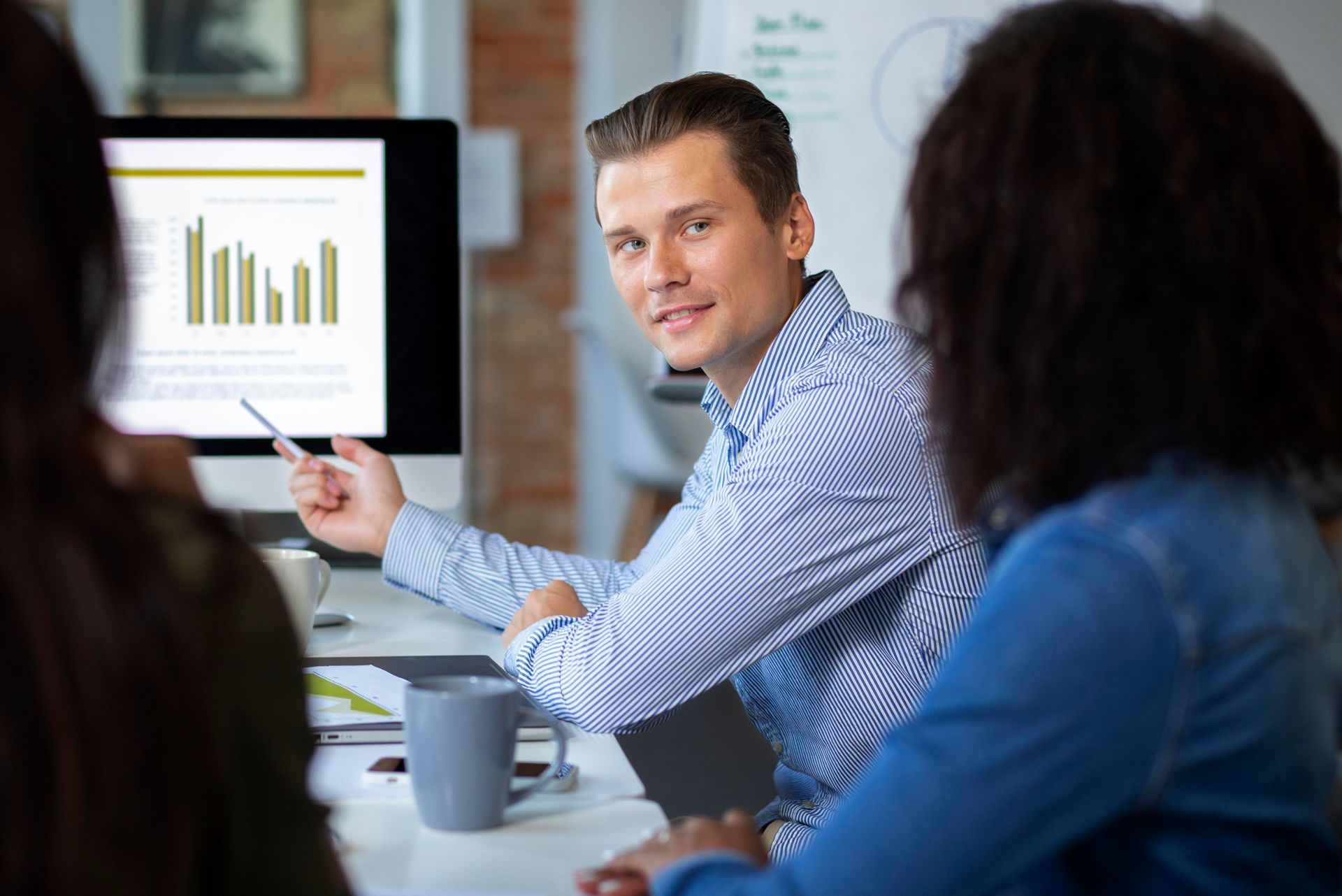 A business professional in a striped shirt pointing at a data chart on a monitor while engaged in a collaborative discussion with colleagues, symbolizing the economic shift toward group captive insurance models.