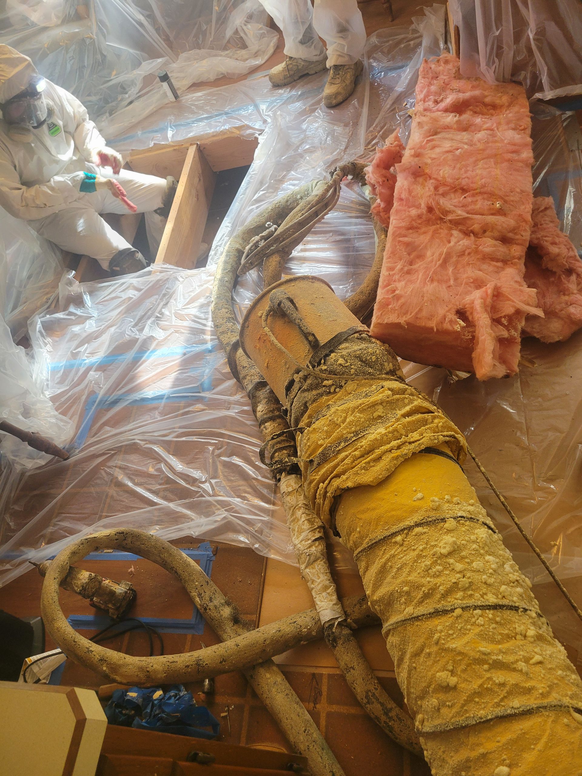 Workers in protective suits removing asbestos from pipes and insulation in a confined space.