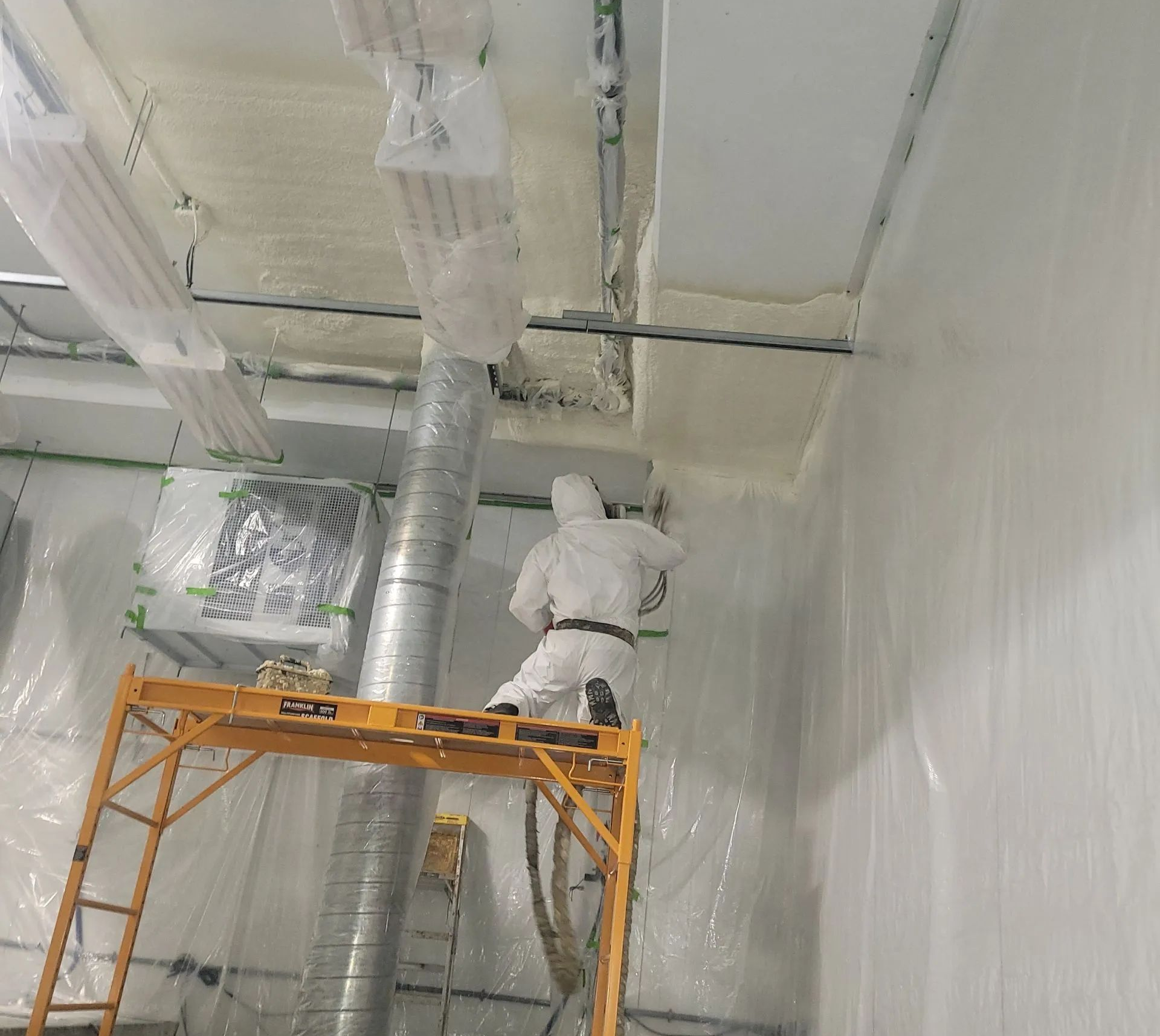 Person in protective suit spraying foam insulation on a ceiling, standing on a scaffolding inside a building.