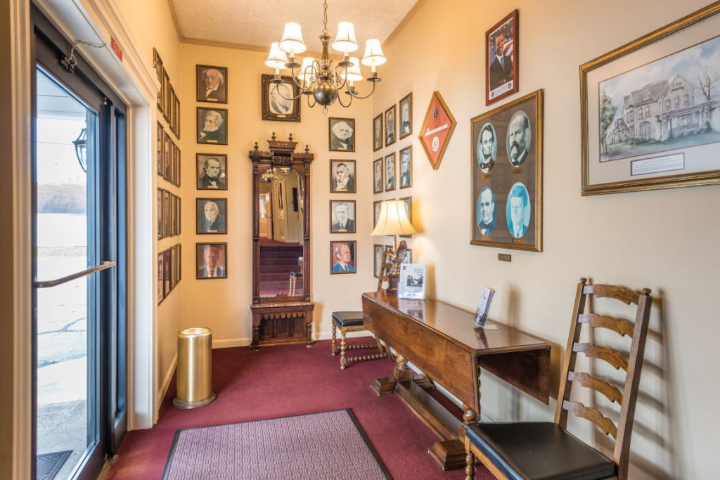 Narrow hallway with red carpet, wooden benches, framed art, and a chandelier-lit ceiling.