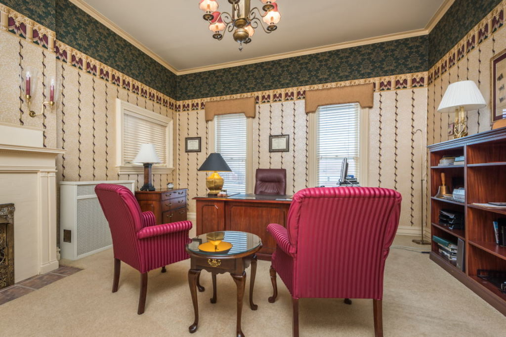 Cozy living room with red armchairs, wooden table, fireplace, bookshelves, and windows with lace curtains