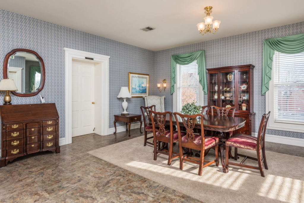 Dining room with wooden table, china cabinet, striped wallpaper, and green drapes by bright windows.