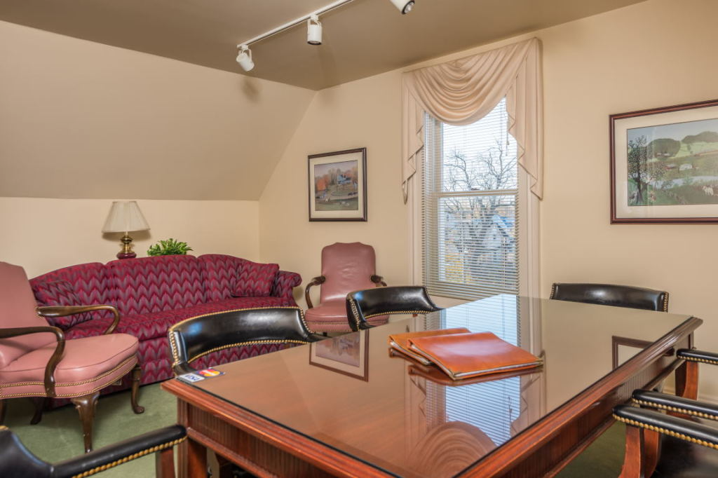 Conference room with polished wooden table, pink chairs, sofa, lamp, and curtained window