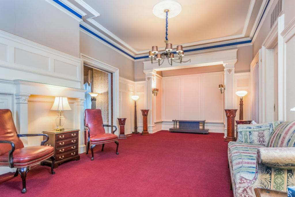 Elegant room with red carpet, chandeliers, pink walls, and upholstered chairs by a fireplace.