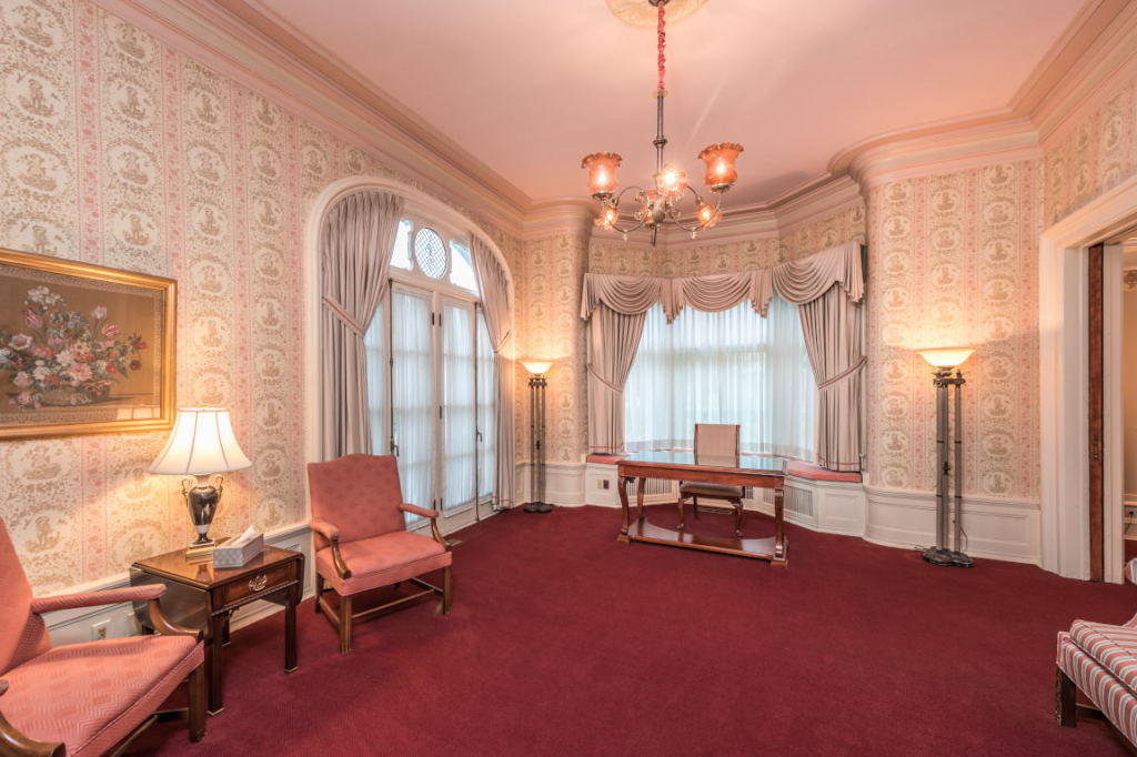 Elegant pink Victorian-style room with ornate wallpaper, chandeliers, and red carpet, furnished with chairs and table