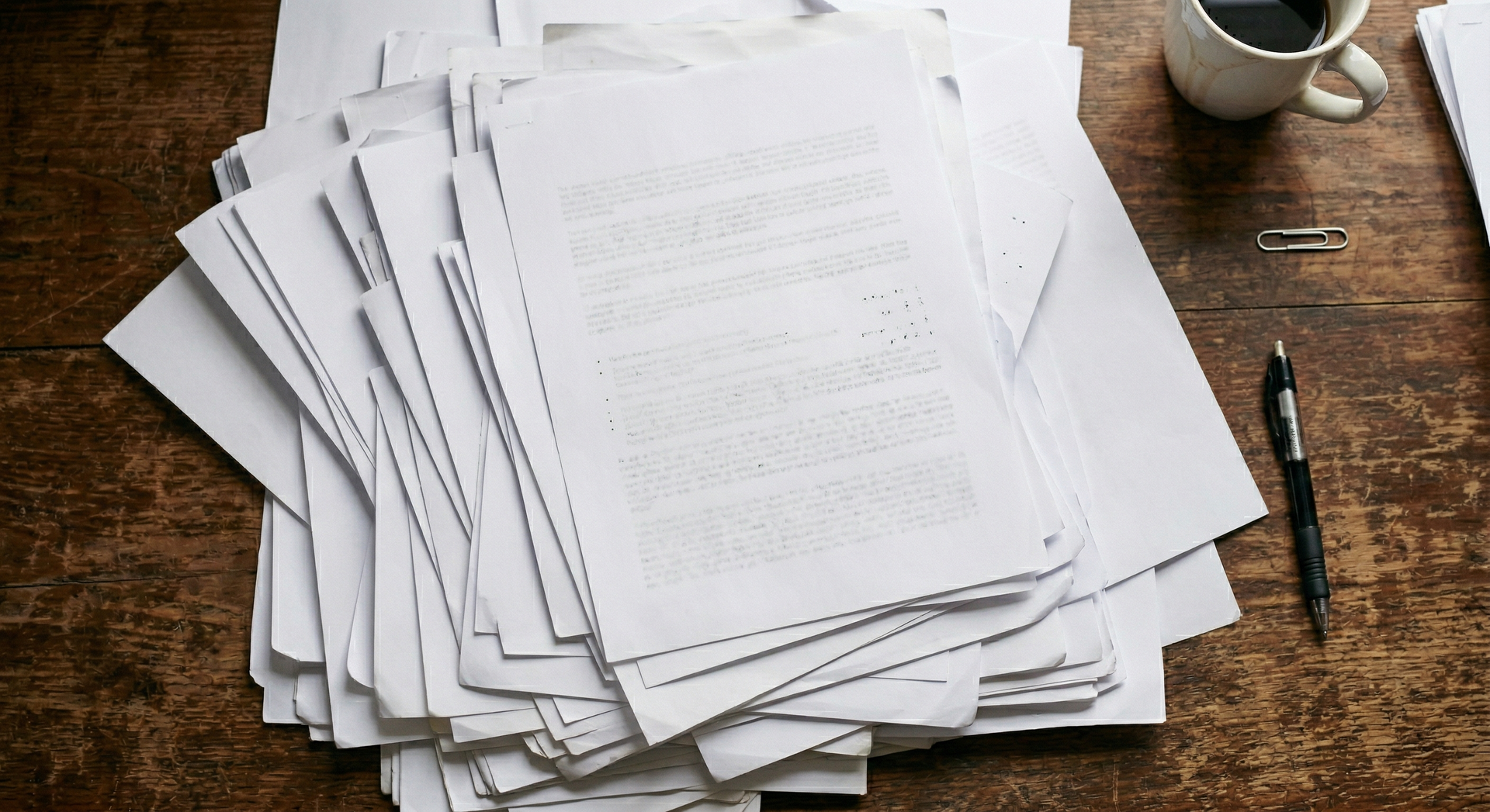 Stack of papers on a wooden desk with a coffee cup and pen nearby.