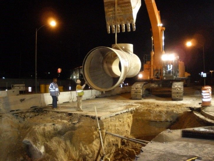 Night road construction with excavator lifting a large concrete pipe, workers nearby