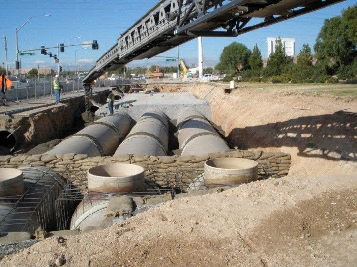 Construction site with large drainage pipes in a trench beside a road, under a crane arm