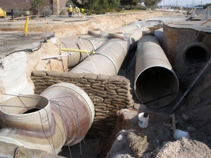 Large metal pipes laid in a trench at a construction site, with sandbags and excavation equipment nearby
