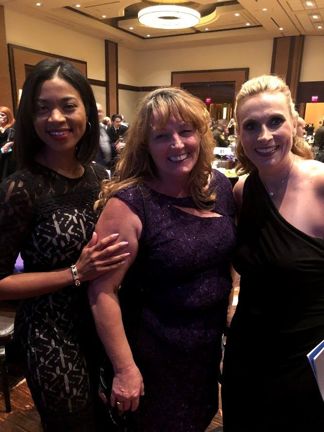 Three people posing together indoors, smiling at a formal event.
