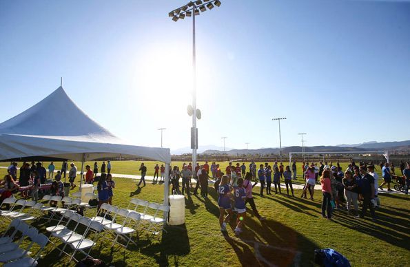 Crowd gathered on a sunny sports field beside a white tent and rows of chairs, with stadium lights overhead