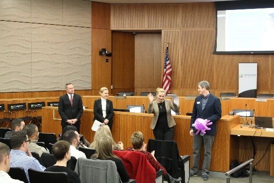 People standing at the front of a meeting room, one holding a purple gift bag, with an American flag behind them