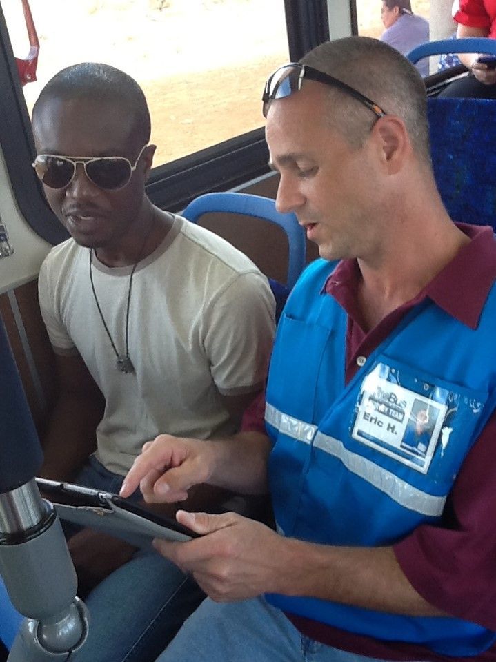 Two men seated on a bus, one showing the other something on a tablet.