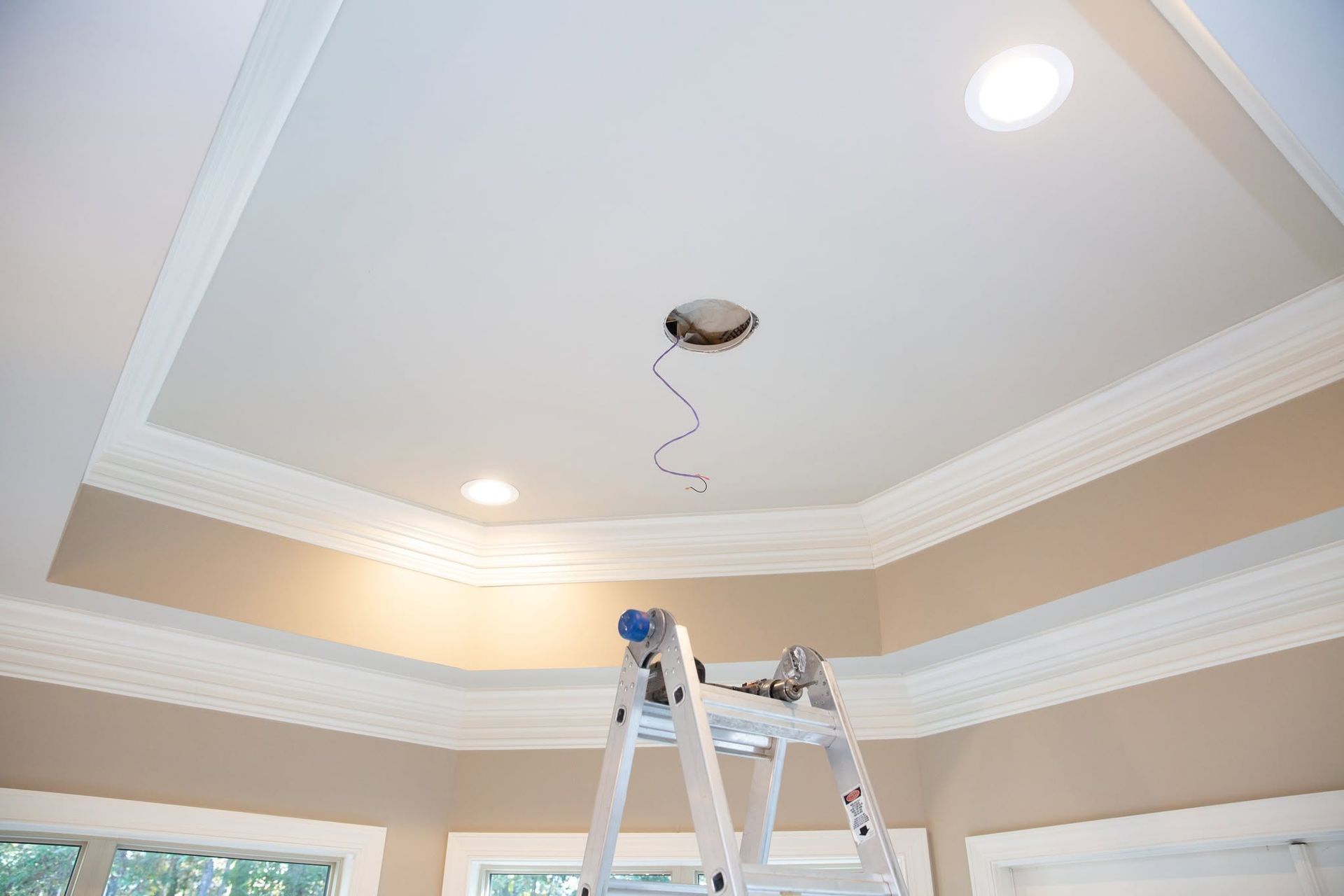 Ceiling under renovation with recessed lights, crown molding, and a ladder in a room with beige walls.