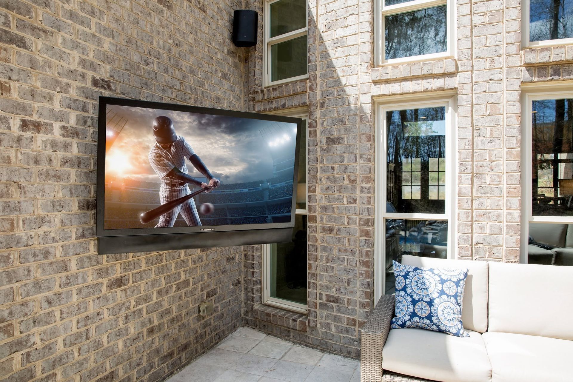 Wall-mounted TV displaying a surfer on a stone patio beside a white sofa and windows