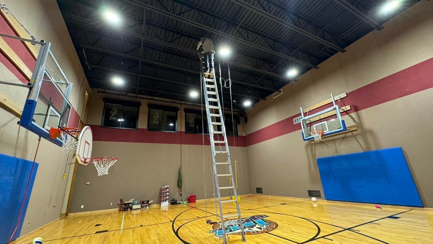 Indoor gym with a tall ladder centered under a high ceiling, basketball hoops, and blue wall mats.