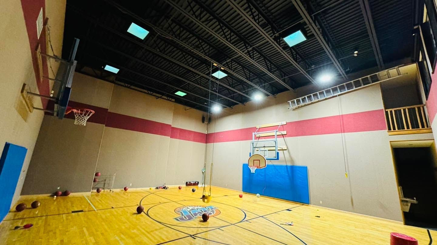 Indoor basketball court with polished wood floor, blue wall padding, and high ceiling lights