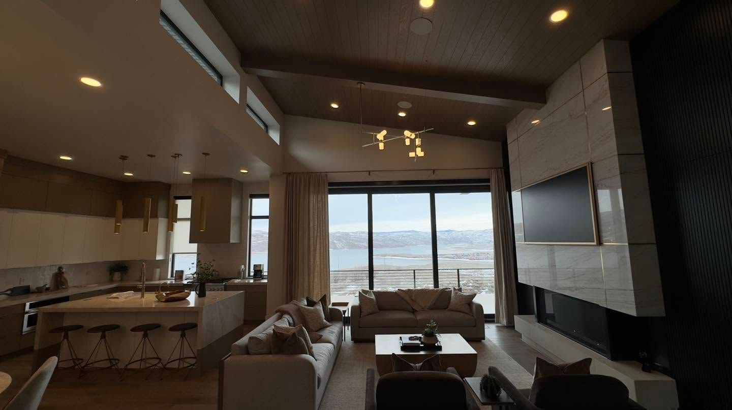 Modern living room with large windows, sofa seating, kitchen island, and a wall-mounted TV overlooking a city view